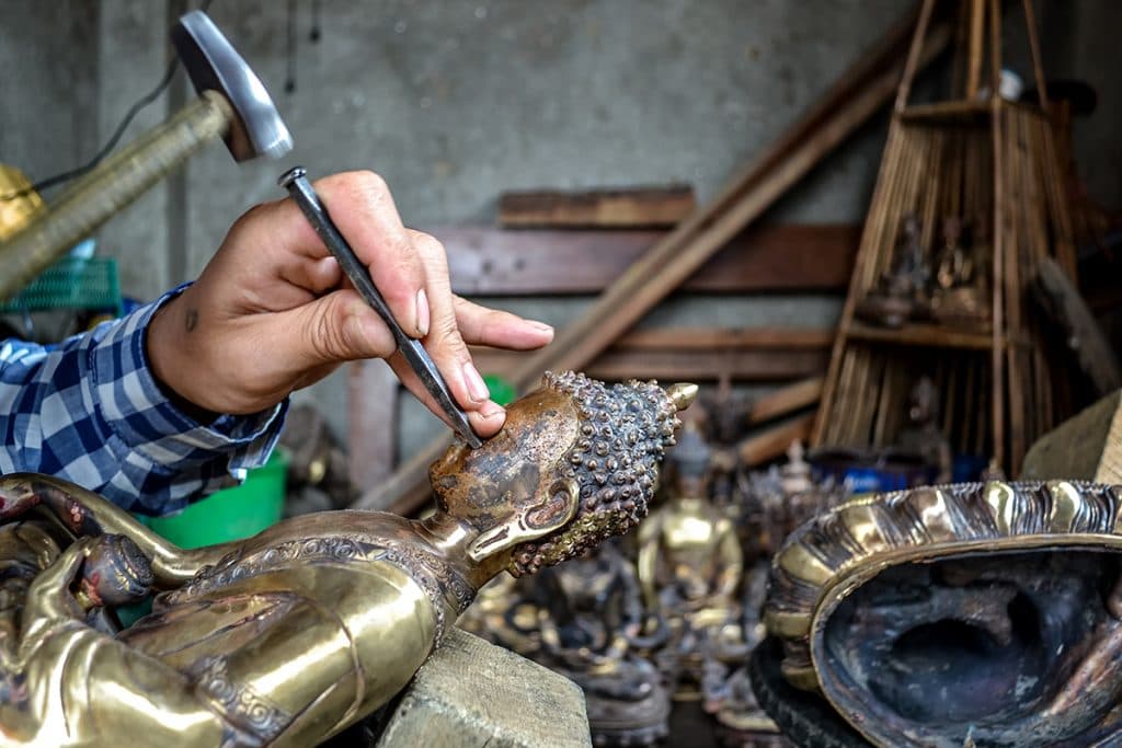 An artisan’s hands using a hammer and chisel to detail a bronze Buddha in a Patan, Nepal workshop
